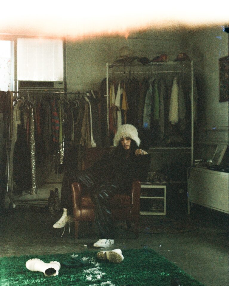 Person in fluffy hat sits in armchair surrounded by clothes.